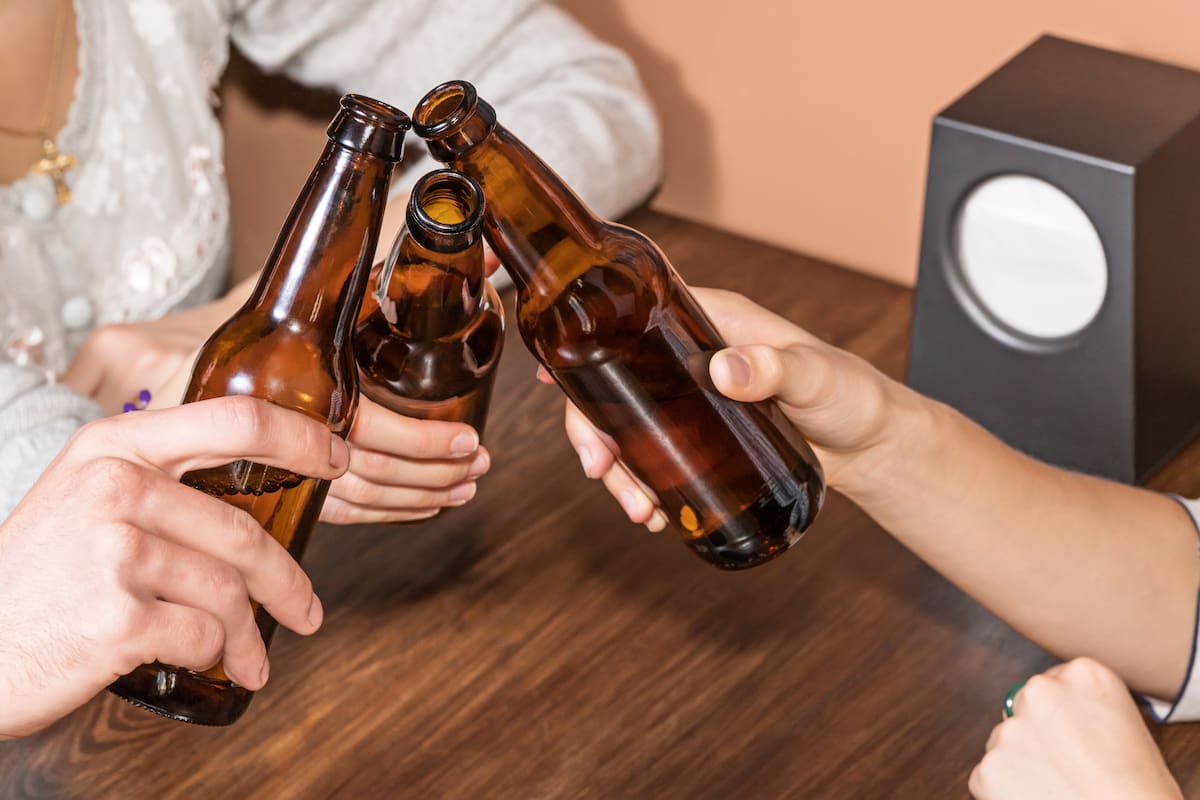 Three friends sitting at a table in a bar with beer bottles clink, closeup