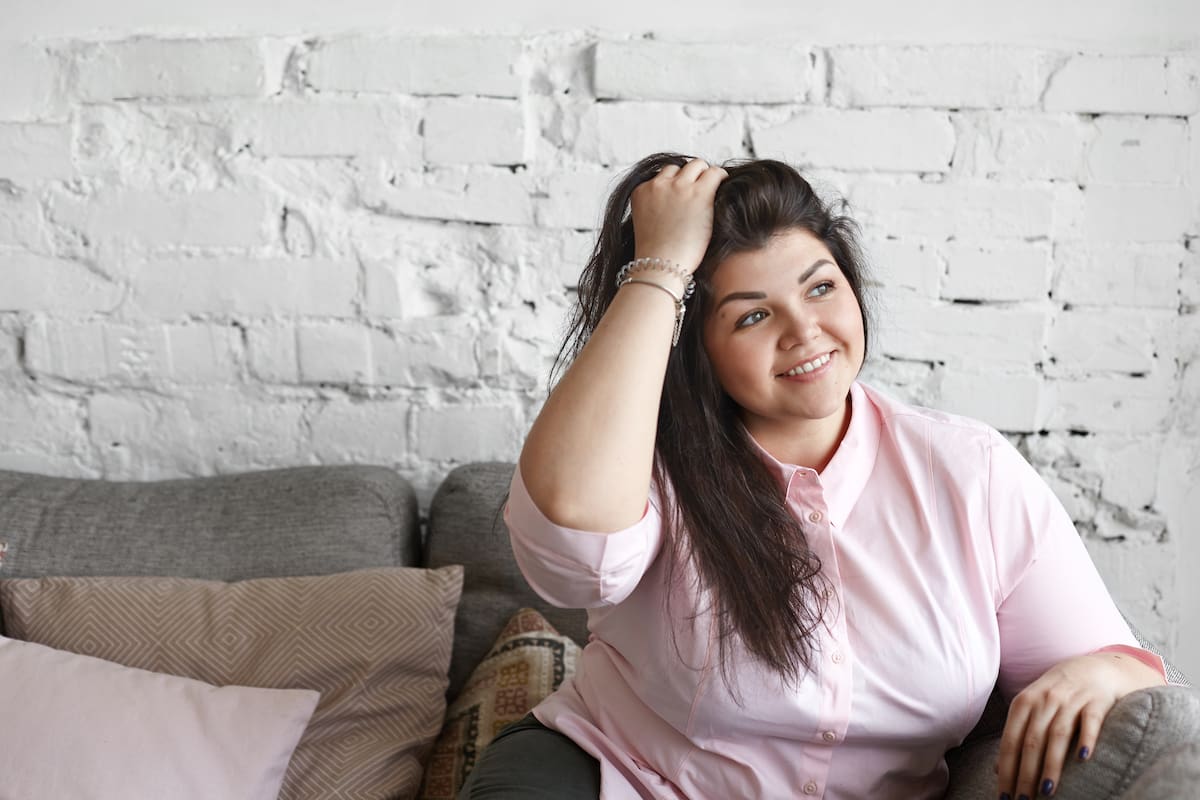 People and lifestyle concept. Body positivity. Indoor portrait of positive young Caucasian female with chubby cheeks enjoying nice day at home, smiling happily, touching her long dark hair