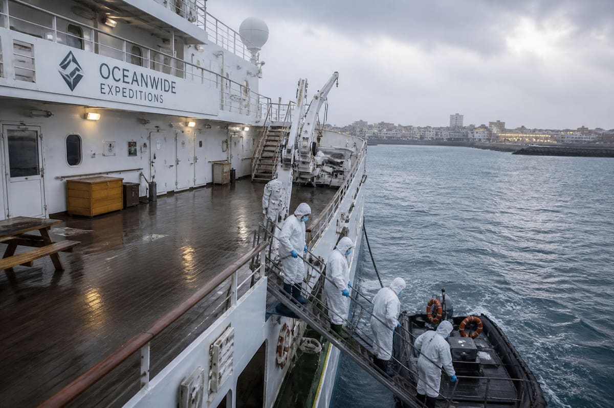 Representación gráfica de las cubiertas del buque, Oceanwide Expeditions aparecen casi completamente abandonadas y solo se observa el movimiento ocasional de personas con trajes de protección completos, mamelucos blancos, botas y mascarillas, recordando las imágenes que se veían durante la pandemia del Coronavirus.